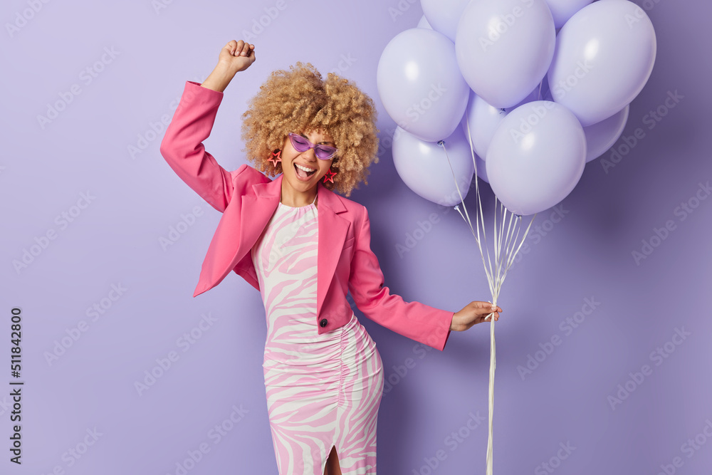 Cheerful curly haired beautiful woman dances with rhythm of music dressed in festive clothes enjoys special occasion holds bunch of balloons isolated over purple background. Party time concept Cheerful curly haired beautiful woman dances with rhythm of music dressed in festive clothes enjoys special occasion holds bunch of balloons isolated over purple background. Party time concept