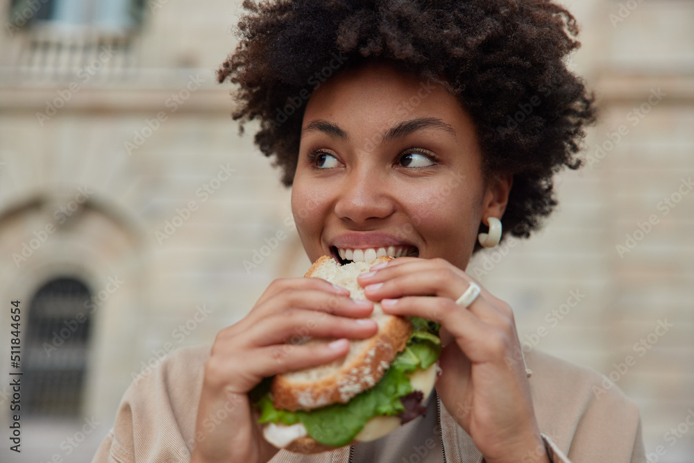 © wayhome.studio  - Pretty curly haired woman bites delicious sandwich poses outdoors at street looks away dressed casually has quick snack while walking outside being hungry. People lifestyle and fast food concept