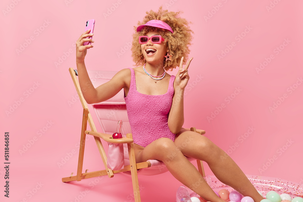Beautiful cheerful woman takes selfie on smartphone makes peace gesture wears sunglasses and bathingsuit has fun poses on comfortable deck chair isolated over pink background. Summer time concept Beautiful cheerful woman takes selfie on smartphone makes peace gesture wears sunglasses and bathingsuit has fun poses on comfortable deck chair isolated over pink background. Summer time concept
