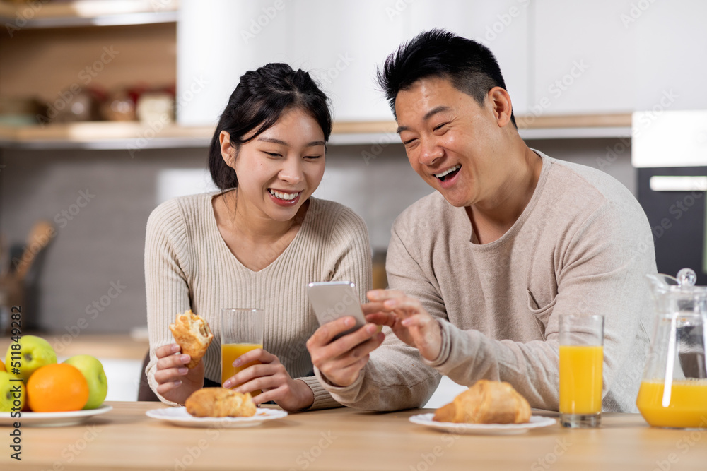 Happy chinese couple using smartphone while having breakfast