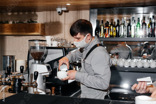 A masked barista prepares an exquisite delicious coffee at the bar in a coffee shop. The work of restaurants and cafes during the pandemic.