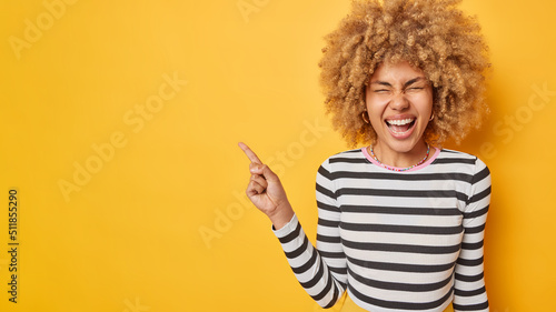 Fotografiet Overjoyed happy young woman with curly blonde hair points away on blank space advertises something laughs gladfully wears casual striped jumper isolated over vivid yellow background