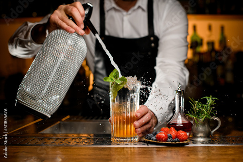 Close-up view of glass with cocktail and sprig of mint in which bartender adds liquid from siphon photo
