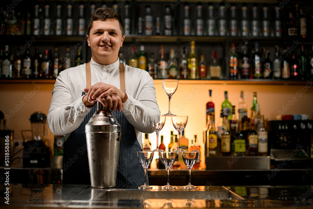 smiling man bartender holds large shaker and pyramid of empty glasses stands nearby on bar