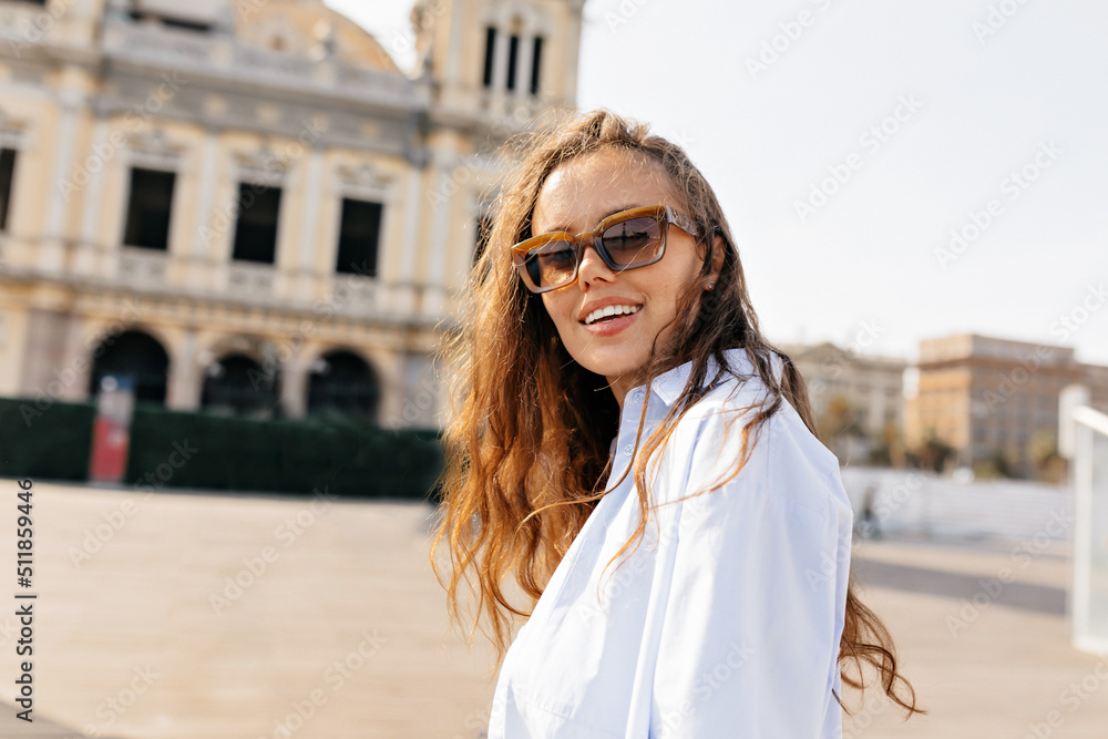 Fototapeta premium Happy smiling girl with loose wavy hair wearing light summer shirt is posing at camera in sunlight with happy emotions in the city