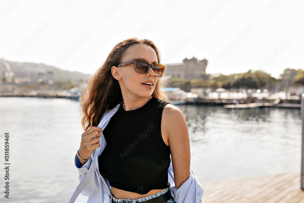 Attractive lovely girl in black top and blue shirt wearing sunglasses looking aside with smile and enjoying walking outside in sunny warm day