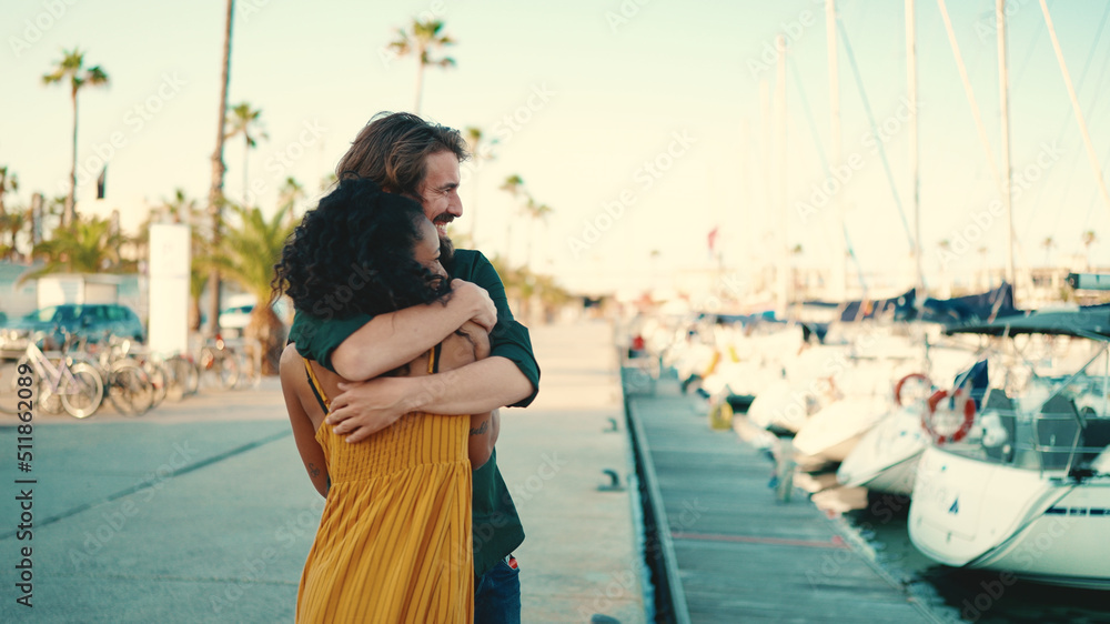 Closeup, happy meeting of young interracial couple. Young man and woman joyfully embrace when they meet on the embankment on the yacht background. Camera moves around people
