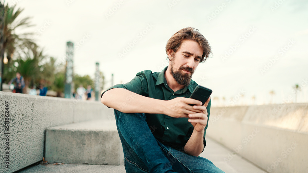 Fototapeta premium Close-up portrait of a man with a beard recording a voice message on the embankment background. Closeup of a young hipster male using a mobile phone