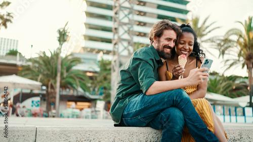 portrait of happy man and smiling woman watching video on smartphone. Close-up of joyful young interracial couple browsing photos on mobile phone on urban city background