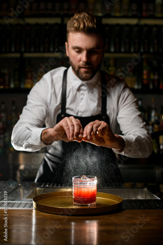 professional man bartender skillfully sprinkles by citrus peel on glass with cocktail