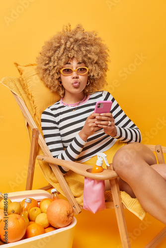 Lovely curly haired young woman keeps lips folded has romantic mood surfs internet on mobile phone wears striped jumper and sunglasses sits on comfortable deck chair against yellow background