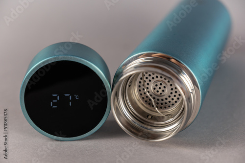 Thermos with open lid on gray background. Cylindrical lid with LED digital display. Temperature reading 27 degrees Celsius. Vacuum flask of turquoise color. Equipment for sports, hiking, travel