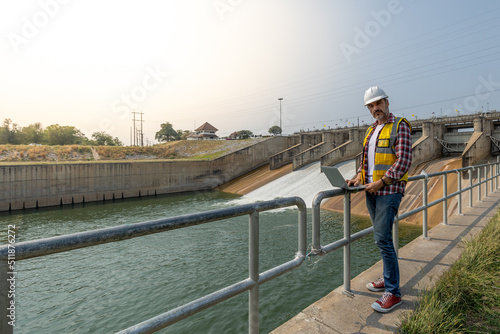 A dam engineering doing his checking routine. He is wearing a white hard hat and yellow transparent vest. He is standing by the rail by the dam.