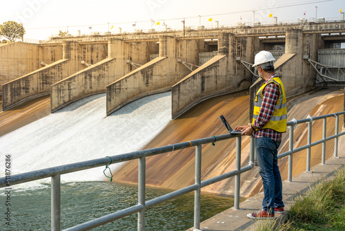 A dam engineering doing his checking routine. He is wearing a white hard hat and yellow transparent vest. He is standing by the rail by the dam. photo
