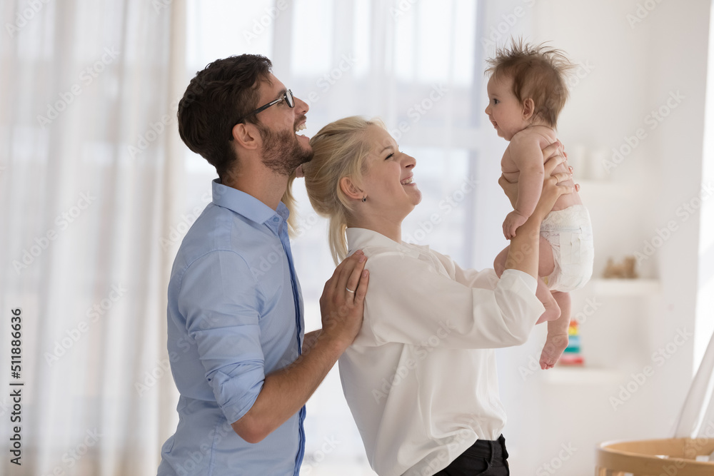 Obraz premium Overjoyed young couple enjoy playtime, spend leisure standing in modern nursery play with their baby cute adorable infant in diaper. Family bond, unconditional love, happy parenthood, babyhood concept