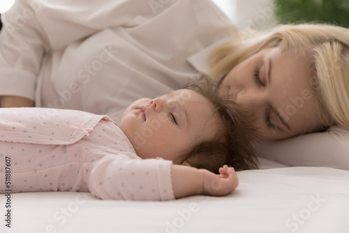 Close up shot peaceful newborn baby and young mom sleeping in bed, lying down with eyes closed on fresh white sheets looking carefree rest together at home. Sweet dreams, protection, maternity concept