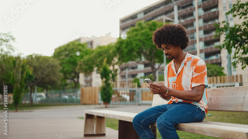 Close-up of young African American man wearing shirt sitting on park bench using his smartphone. Closeup of smiling african american man looking at photo video on mobile phone. Lifestyle concept © Andrii Nekrasov