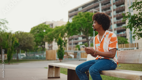 Close-up of young African American man wearing shirt sitting on park bench using his smartphone. Closeup of smiling african american man looking at photo video on mobile phone. Lifestyle concept