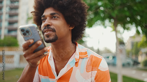 Close-up porttrait of young African American man wearing shirt sitting on park bench using his smartphone. Smiling man sends voice message on mobile phone.
