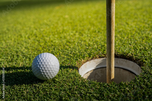 Choose focus. A white golf ball near the golf hole and a flagpole on green grass.