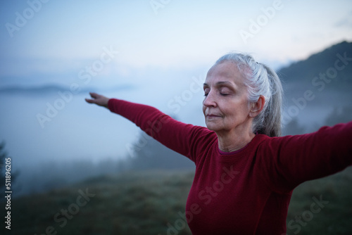 Senior woman doing breathing exercise in nature on early morning with fog and mountains in background.