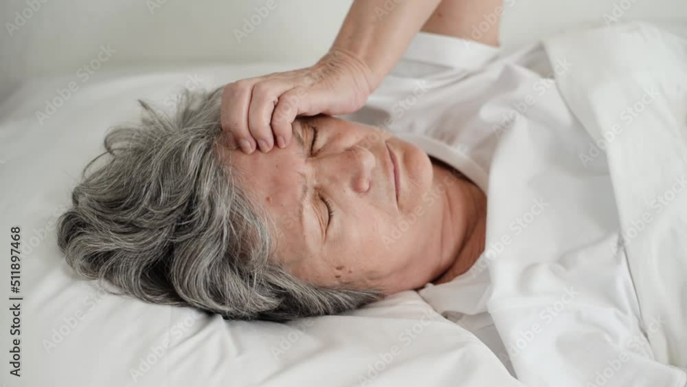 Close-up tired senior woman holding her head while lying with eyes closed on bed in bedroom. Headache, fatigue, insomnia concept Close-up tired senior woman holding her head while lying with eyes closed on bed in bedroom. Headache, fatigue, insomnia concept
