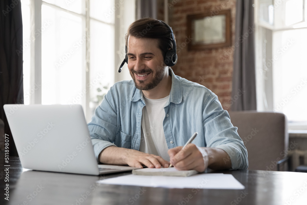 Smiling millennial man wear headset take part in educational webinar, on-line lesson, remote effective english course makes notes sit at desk with laptop. Virtual meeting, tuition, e-learning concept - obrazy, fototapety, plakaty 