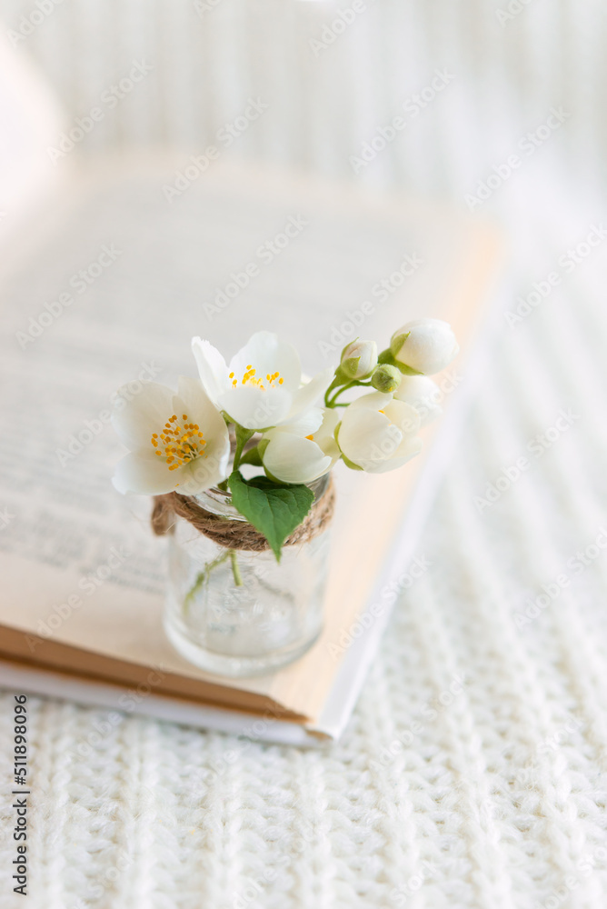 Close-up of a bouquet of white flowers in a blurred glass vase on an open book on a white knitted background. Slow life concept. Close-up of a bouquet of white flowers in a blurred glass vase on an open book on a white knitted background. Slow life concept.