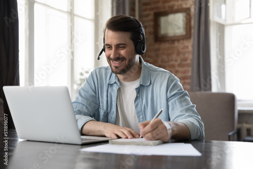 Smiling millennial man wear headset take part in educational webinar, on-line lesson, remote effective english course makes notes sit at desk with laptop. Virtual meeting, tuition, e-learning concept