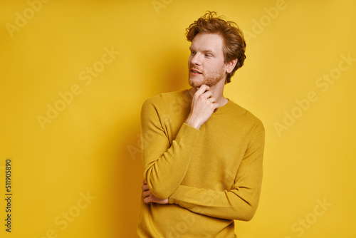 Handsome redhead man holding hand on chin while standing against yellow background