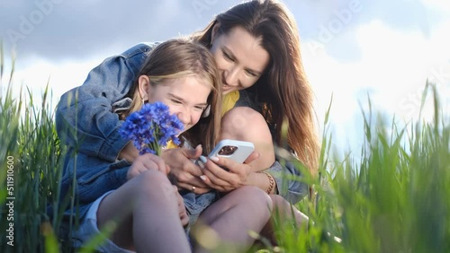 Young mother and daughter relaxing in nature in the park, they have fun laughing using a smartphone photo