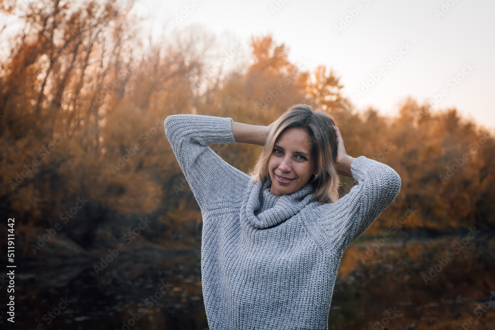 Close-up of charming blonde woman standing in forest, looking at camera smiling with both hands behind head with trees covered with sunset in background. Autumn walk in forest. Close-up of charming blonde woman standing in forest, looking at camera smiling with both hands behind head with trees covered with sunset in background. Autumn walk in forest.