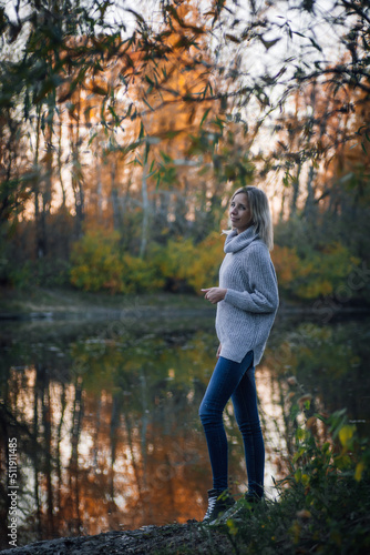 Beautiful blonde woman standing by river in forest, looking at camera and posing with trees in background being part of nature, wearing blue jeans and grey sweater. Autumn walk in forest.