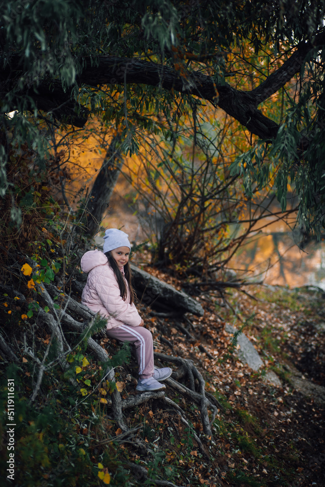 Naklejka premium Young Caucasian girl sitting on tree roots in forest, smiling, looking at camera near river with willow trees in background, wearing hat, pink jacket, sports trousers, sneakers. Autumn walk in forest.