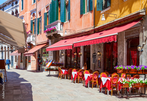 Old street with colorful facades and red tables of cafe at a sunny morning, Venice, Italy.