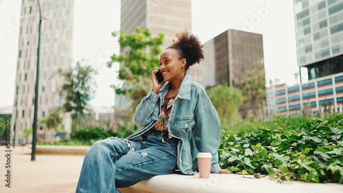 Closeup portret cute African girl with ponytail, wearing denim jacket, sits with morning coffee and makes stream, video call on modern city background.