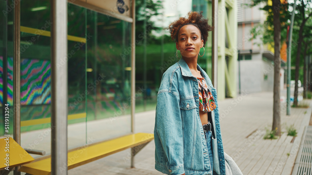 Cute African girl with ponytail, wearing denim jacket, in crop top with national pattern, standing at the bus stop and smiling Cute African girl with ponytail, wearing denim jacket, in crop top with national pattern, standing at the bus stop and smiling