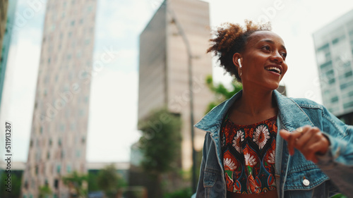 Closeup  smiling African girl with ponytail wearing denim jacket  in crop top with national pattern listening to music on headphones and dancing outdoors.