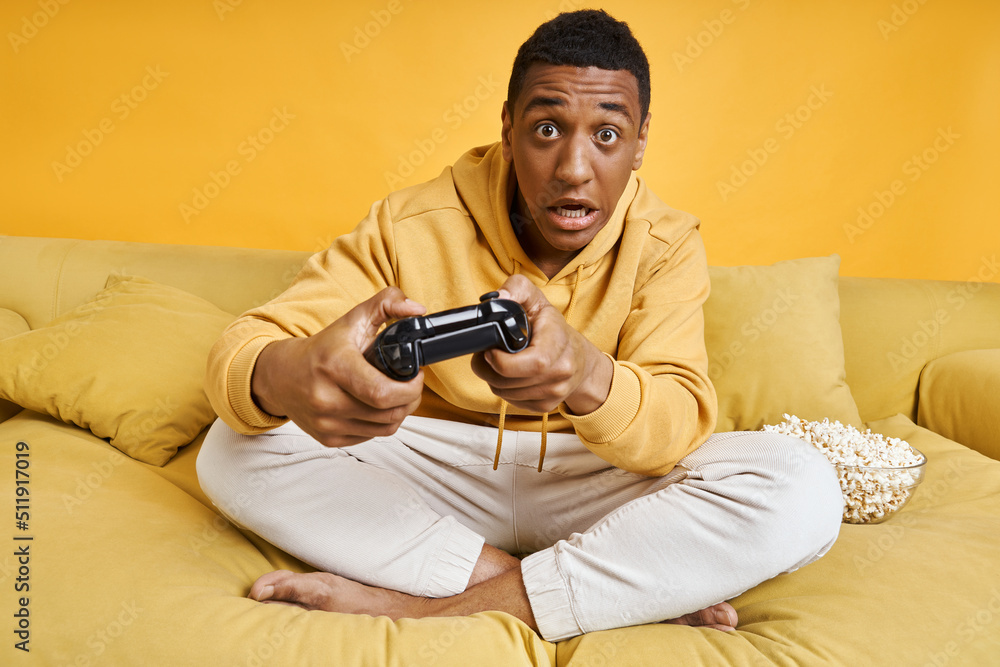 Worried young man playing video games while sitting on the couch with yellow background Worried young man playing video games while sitting on the couch with yellow background