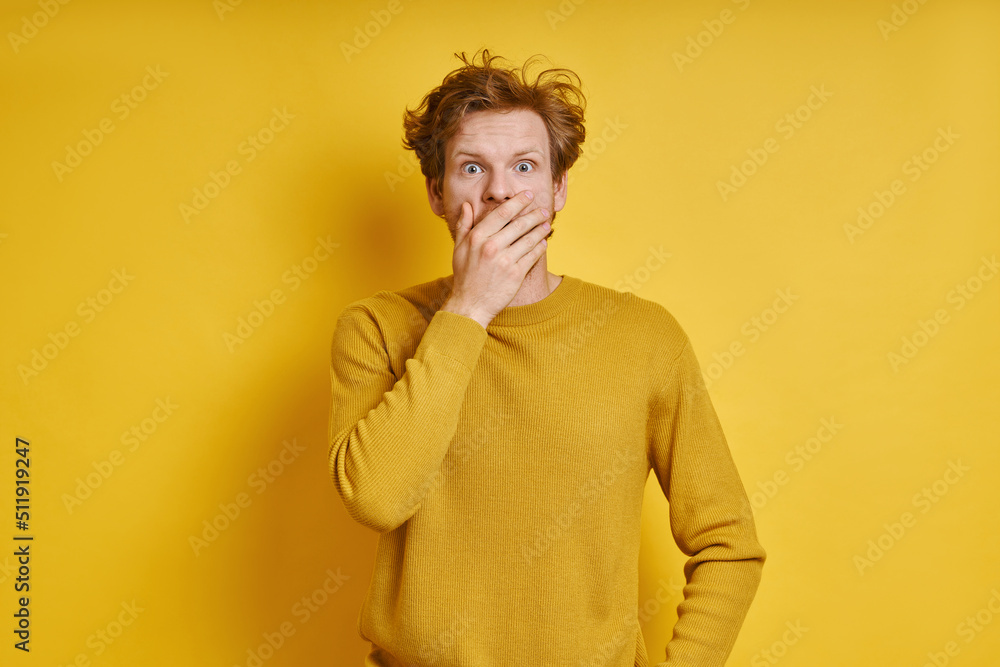 Shocked redhead man covering mouth with hand while standing against yellow background