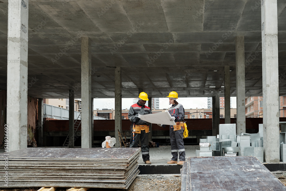 Two modern intercultural builders in workwear discussing architectural plan while holding unrolled blueprint at construction area Two modern intercultural builders in workwear discussing architectural plan while holding unrolled blueprint at construction area