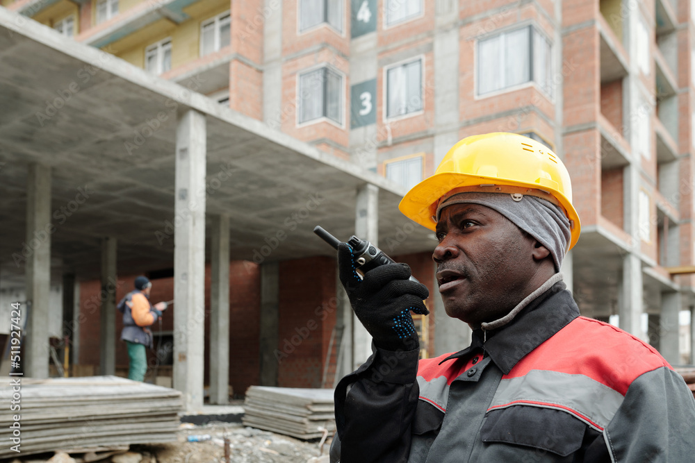 Aged builder in hardhat and workwear speaking in walkie-talkie while working at construction site against unfinished concrete structure Aged builder in hardhat and workwear speaking in walkie-talkie while working at construction site against unfinished concrete structure