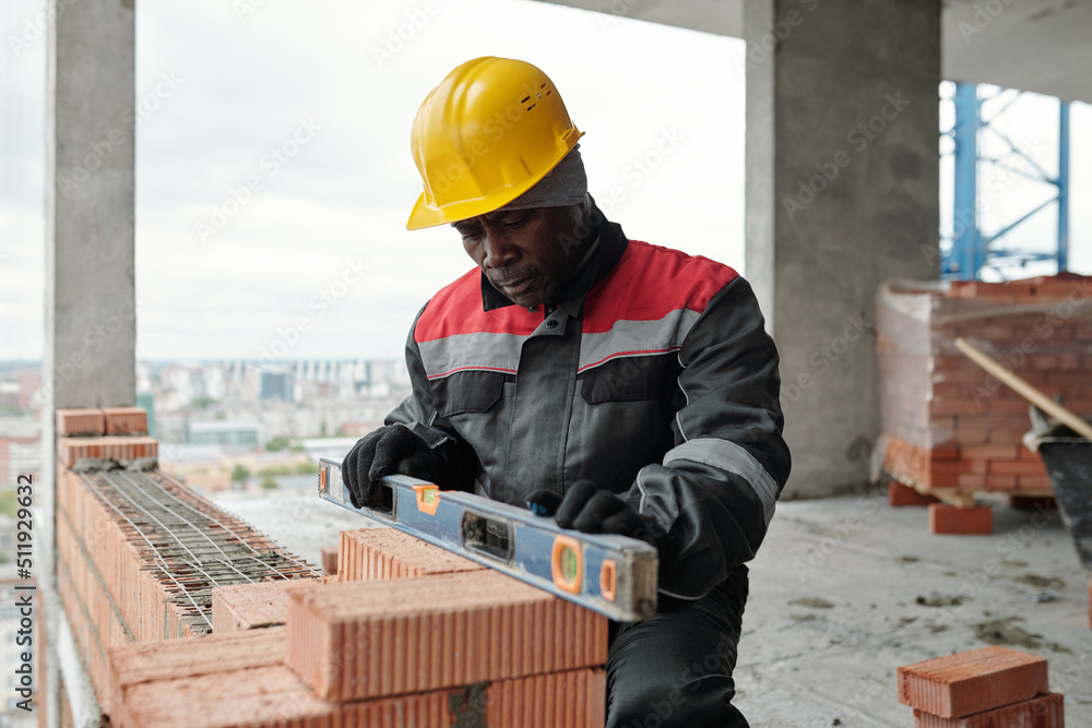 Mature builder in workwear and protective helmet keeping level handtool on surgace of brick layer while checking flatness of wall Mature builder in workwear and protective helmet keeping level handtool on surgace of brick layer while checking flatness of wall