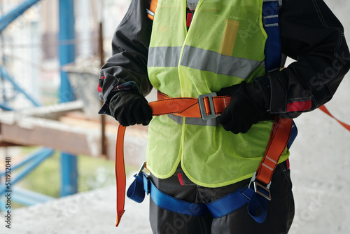 Young builder in gloves and reflective vest fastening safety belt on waist while preparing for building work at construction site