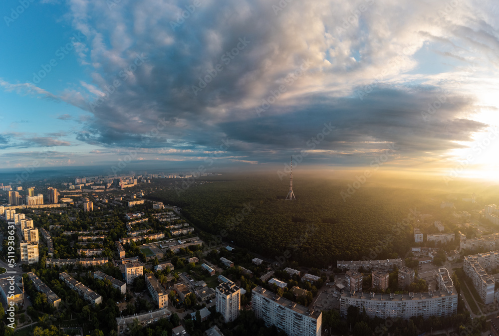 Aerial panorama view on sunrise morning city with telecommunication tower in forest near residential district with scenic sun shining in cloudy sky. Kharkiv, Ukraine