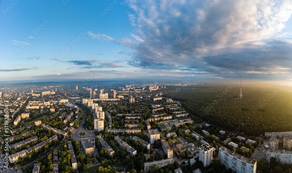 Aerial panorama view on sunrise morning city with telecommunication tower in forest near residential district with scenic sun shining in cloudy sky. Pavlovo Pole, Kharkiv Ukraine