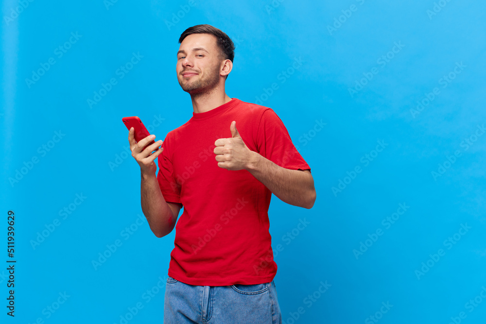 Enjoyed smiling tanned handsome man in red t-shirt doing online shopping with phone show thumb up posing isolated on blue studio background. Copy space Banner Mockup. Online People lifestyle concept