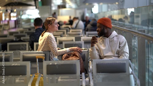 New acquaintances during flight delay. Outgoing African American hipster male with coffee and pretty Caucasian woman traveler talk about different topics sitting on chairs at terminal. Ttravel concept photo