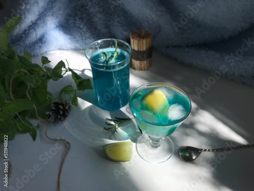 Blue cold drink in a glass with ice cubes on white desk with blue background. Also used mint leaves and rosemary, rope, spoon with natual sunlight from the open window