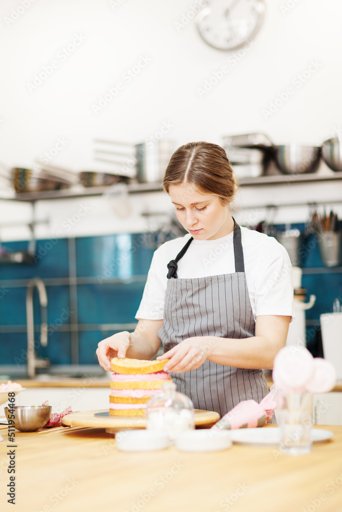 Female pastry chef making delicious cake. Woman stacking sponge layers with pink cream between them in bakery kitchen Female pastry chef making delicious cake. Woman stacking sponge layers with pink cream between them in bakery kitchen